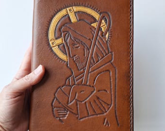 May include: A brown leather book cover with a religious image embossed on the front. The image depicts a figure with a halo, holding a staff and a lamb. The cover is stitched along the edges.