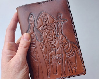 May include: A brown leather wallet with a tooled design of a person wearing a mitre and robe. The design is embossed into the leather.