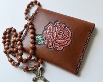 May include: A brown leather wallet with a carved pink rose design and a wooden rosary with a silver crucifix. The wallet is rectangular and has a stitched border. The rosary beads are a dark brown color.