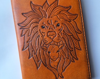 May include: A tan leather notebook cover featuring a detailed embossed design of a lion with a full mane and a small lamb nestled within the lion's mane. The cover has visible dark brown stitching along the edges and a thin black bookmark ribbon.
