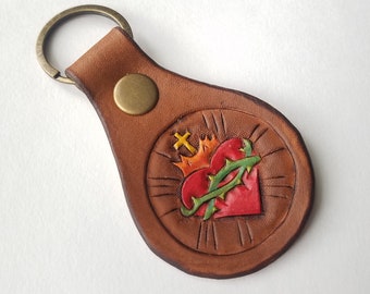 May include: A brown leather keychain with a circular design featuring a red heart with a crown and thorns. The heart is surrounded by a gold halo and a green vine.