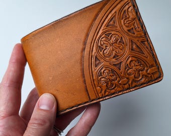 May include: A brown leather wallet with a decorative embossed design on the front. The wallet is held in a hand and has a classic bi-fold design. The embossed pattern features floral and geometric shapes. The leather has a natural, aged appearance.