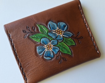 May include: Brown leather wallet with hand-painted blue flowers and green leaves. The floral design is embossed on the front of the wallet. The edges are stitched with a contrasting thread.