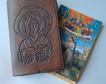 May include: A brown leather cover featuring a hand-tooled embossed image of a figure with hands outstretched, positioned next to a Magnificat December 2022 Vol. 24, No. 10 booklet with a colorful religious illustration on its front cover.