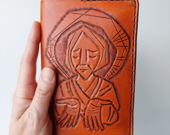 May include: A brown leather wallet with a tooled design of a woman with her hands clasped in prayer. The woman is wearing a hooded robe.