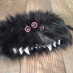 K&ouml;nnte beinhalten: Schwarzes, flauschiges Monster mit drei rosa und schwarzen Augen und wei&szlig;en Z&auml;hnen. Das Monster hat einen schwarzen Riemen, der an der R&uuml;ckseite befestigt ist.