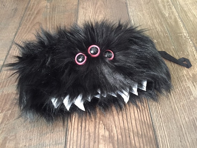 K&ouml;nnte beinhalten: Schwarzes, flauschiges Monster mit drei rosa und schwarzen Augen und wei&szlig;en Z&auml;hnen. Das Monster hat einen schwarzen Riemen, der an der R&uuml;ckseite befestigt ist.