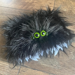 May include: Black fuzzy spider plush toy with three green googly eyes. The spider has a white, jagged edge around the bottom.
