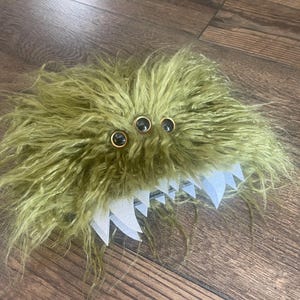 May include: A whimsical, olive green monster hat with three large, black-rimmed eyes and a row of white, jagged teeth. The hat is made of shaggy, textured material, creating a playful and unique accessory.