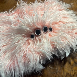 May include: A plush, pink and white furry creature with large, black, googly eyes. The creature has a fluffy, shaggy texture and a whimsical appearance. The eyes are arranged in a cluster, giving it a playful look. The overall design suggests a handmade toy or decorative item.
