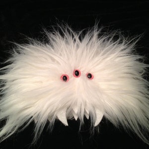 May include: A white fluffy creature with three pink-rimmed black eyes. The creature has a toothy grin.
