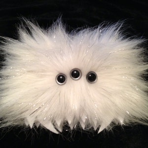 May include: A white fluffy toy with three black eyes. The toy has a spiky, jagged bottom edge.