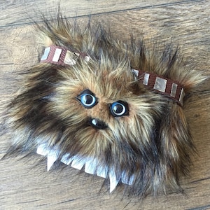 May include: A brown and black furry Chewbacca-inspired plush toy with blue eyes and a brown leather strap with silver accents.