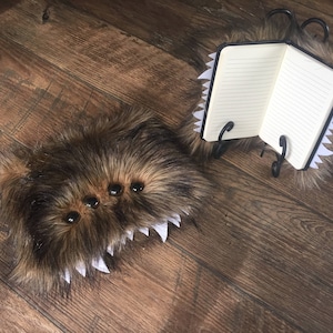 May include: A quirky notebook with a brown, furry cover, four black eye-like details, and white, jagged teeth. The notebook is open, displaying lined pages, and is supported by a black metal stand. A matching closed notebook is also present.