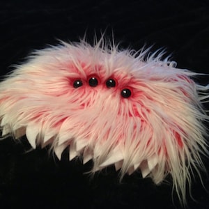 May include: A pink and white fuzzy creature with three black eyes and a jagged mouth.
