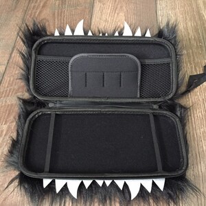 K&ouml;nnte beinhalten: Schwarze und wei&szlig;e, flauschige Tragetasche mit Rei&szlig;verschluss und einem Netzfach im Inneren. Die Tasche hat einen wei&szlig;en Kunststoffzahn-Design am Rand.