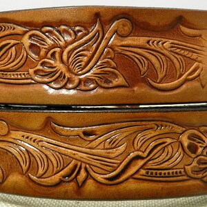 Może przedstawiać: Dwa brązowe skórzane paski z misternymi kwiatowymi zdobieniami. Górny pasek ma wygrawerowane słowo "ST" po prawej stronie.