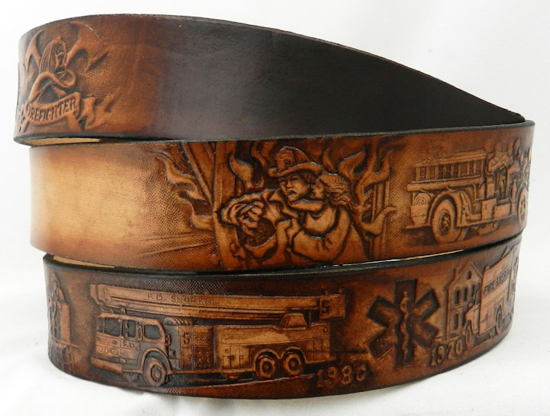 May include: A brown leather belt with a firefighter theme. The belt is decorated with embossed images of firefighters, fire trucks, and flames. The belt also has the text "Firefighter" and the years "1930" and "1970" embossed on it.