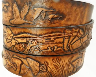 May include: A brown leather belt with three embossed scenes of duck hunting. The top scene shows ducks in flight, the middle scene shows a hunter aiming a shotgun, and the bottom scene shows ducks swimming in a marsh.