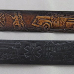 May include: Two leather belts with embossed designs. The top belt is brown with a fire truck and firefighters, and the text "FD" and "1955". The bottom belt is black with a fire truck and the text "Fire Department".