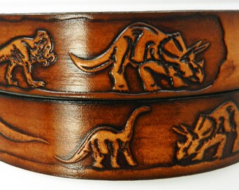 May include: A brown leather belt with dinosaur designs embossed on it. The belt has two layers, and each layer has a different dinosaur design. The top layer features a Tyrannosaurus Rex, a Triceratops, and a Brachiosaurus. The bottom layer features a Stegosaurus and another Triceratops.