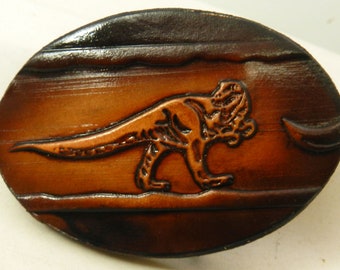 May include: A brown leather belt buckle with a dinosaur design. The dinosaur is embossed in the leather and is facing to the right. The buckle is oval shaped.