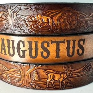 May include: A brown leather belt with a carved design of a deer, a bear, and a tree. The belt has the name "AUGUSTUS" embossed on it.