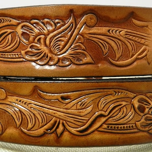 Może przedstawiać: Dwa ułożone na sobie brązowe skórzane paski z misternymi kwiatowymi i rolowanymi wzorami. Górny pasek ma czarny napis "ST". Paski są prawdopodobnie przeznaczone do noszenia w stylu western.