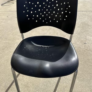 Puede incluir: Silla de plástico negro con respaldo perforado y patas de metal plateado. La silla está sobre una superficie de hormigón.
