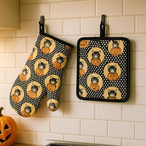 May include: A set of two black and white oven mitts with a Halloween theme. The mitts feature a repeating pattern of black cats holding orange pumpkins. The mitts are hanging on hooks above a stainless steel stove.