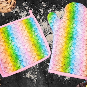 May include: A set of two pink oven mitts with a rainbow mermaid scale pattern. The mitts are made of fabric and have a pink trim.