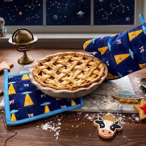 May include: A baked apple pie with a lattice crust is held with a blue oven mitt featuring a UFO and cow design. Matching pot holders and star-shaped cookies are on a wooden surface. A small alien figurine and a globe are in the background.
