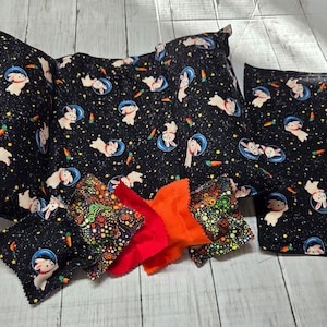 Può includere: Una collezione di accessori per animali domestici con un tessuto nero con una stampa di conigli spaziali. Include un cuscino, un letto rettangolare e diversi fiocchi colorati. I fiocchi sono rossi, arancioni e fantasia.