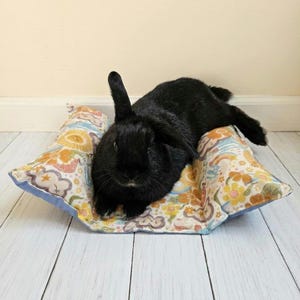 Puede incluir: Un conejo negro sólido descansando sobre una cama rectangular para mascotas. La cama presenta un patrón colorido de flores amarillas, soles naranjas, arcoíris y nubes sobre un fondo blanco, con un borde de tela azul claro en los extremos.