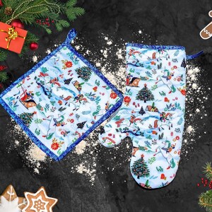 May include: A set of two blue and white Christmas-themed oven mitts. The mitts feature a winter scene with horses, trees, and houses. The larger mitt is a potholder and the smaller mitt is an oven mitt.