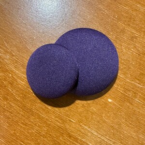 May include: Two purple fabric-covered buttons of different sizes. The larger button is positioned behind the smaller one, creating a layered effect. The buttons have a textured appearance and are set against a wooden surface.