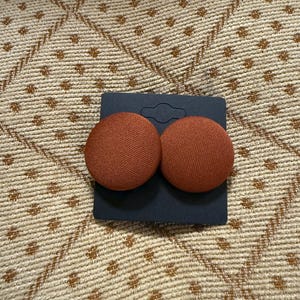 May include: A pair of brown fabric covered button earrings. The buttons are round and have a textured surface.