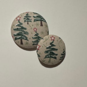 May include: Two round fabric buttons with a beige background and a pattern of green Christmas trees with red stars.