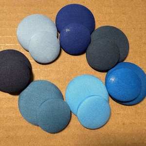 May include: An assortment of fabric-covered buttons in various shades of blue, from light sky blue to deep navy. The buttons are arranged in a circular pattern, showcasing their different sizes and hues. The buttons are on a brown surface.