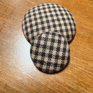 May include: Two circular fabric-covered buttons featuring a black and beige gingham check pattern. The buttons are domed in shape, with one button noticeably larger than the other, both displaying a consistent woven textile texture throughout.