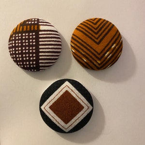 May include: Three round fabric buttons with geometric patterns. The first button has a brown, white, and yellow geometric pattern. The second button has a brown and yellow chevron pattern. The third button has a black background with a brown square surrounded by a white border.
