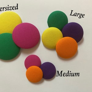 May include: An assortment of colorful, fabric-covered buttons in various sizes: oversized, large, and medium. The buttons are arranged in groups, with colors including yellow, green, pink, orange, and purple. Text labels indicate the size of each group.