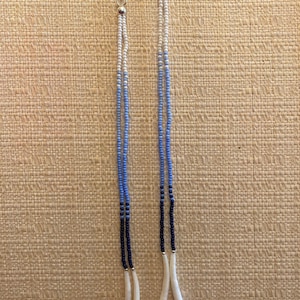 May include: A pair of long, beaded earrings with a gradient of white, light blue, dark blue, and black beads. The earrings have a bone-like pendant at the bottom.