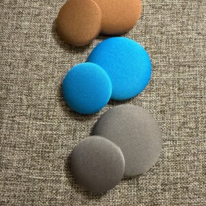 May include: Three sets of round, fabric-covered buttons in varying sizes. The sets are in brown, blue, and gray. The buttons have a textured appearance and are arranged in pairs, with a smaller button next to a larger one.