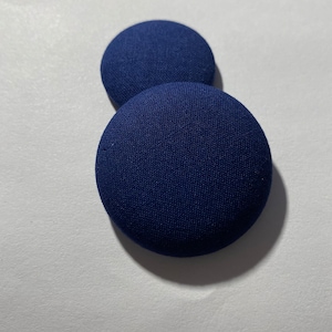 May include: Two navy blue, fabric-covered buttons. The buttons are round and have a slightly raised profile. The fabric has a fine texture, and the buttons are casting a shadow on a white surface. These buttons could be used for sewing or crafts.