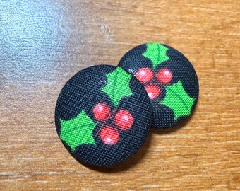 May include: Two round black fabric buttons with a Christmas holly design. Each button features green holly leaves and red berries. The buttons are set against a wooden background.