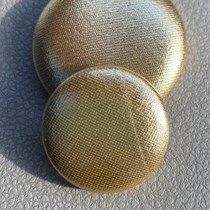 May include: Two gold-colored, fabric-covered buttons of different sizes. The buttons have a textured surface and are set against a gray, textured background. The larger button is approximately 1.5 inches in diameter.