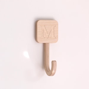 May include: Wooden wall hook with a square top featuring the letter "M". The hook is made of light-coloured wood and has a curved hook shape. The hook is mounted on a white wall.