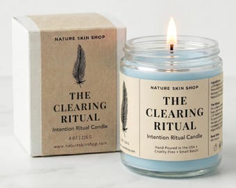 Op de afbeelding: Een lichtblauwe kaars in een doorzichtige glazen pot met een etiket met de tekst The Clearing Ritual, vergezeld van een bijpassende kartonnen doos. De kaars heeft een brandende lont en een veer-afbeelding. Het etiket vermeldt 226 g.