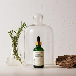 May include: A green glass bottle with a gold dropper, labeled "Follicle Fortifier" sits under a glass cloche. A sprig of rosemary is in a clear glass beaker to the left. The background is white.